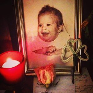 Picture of a baby framed, with a dried flower, lit candle, and a homemade clover in front of it.